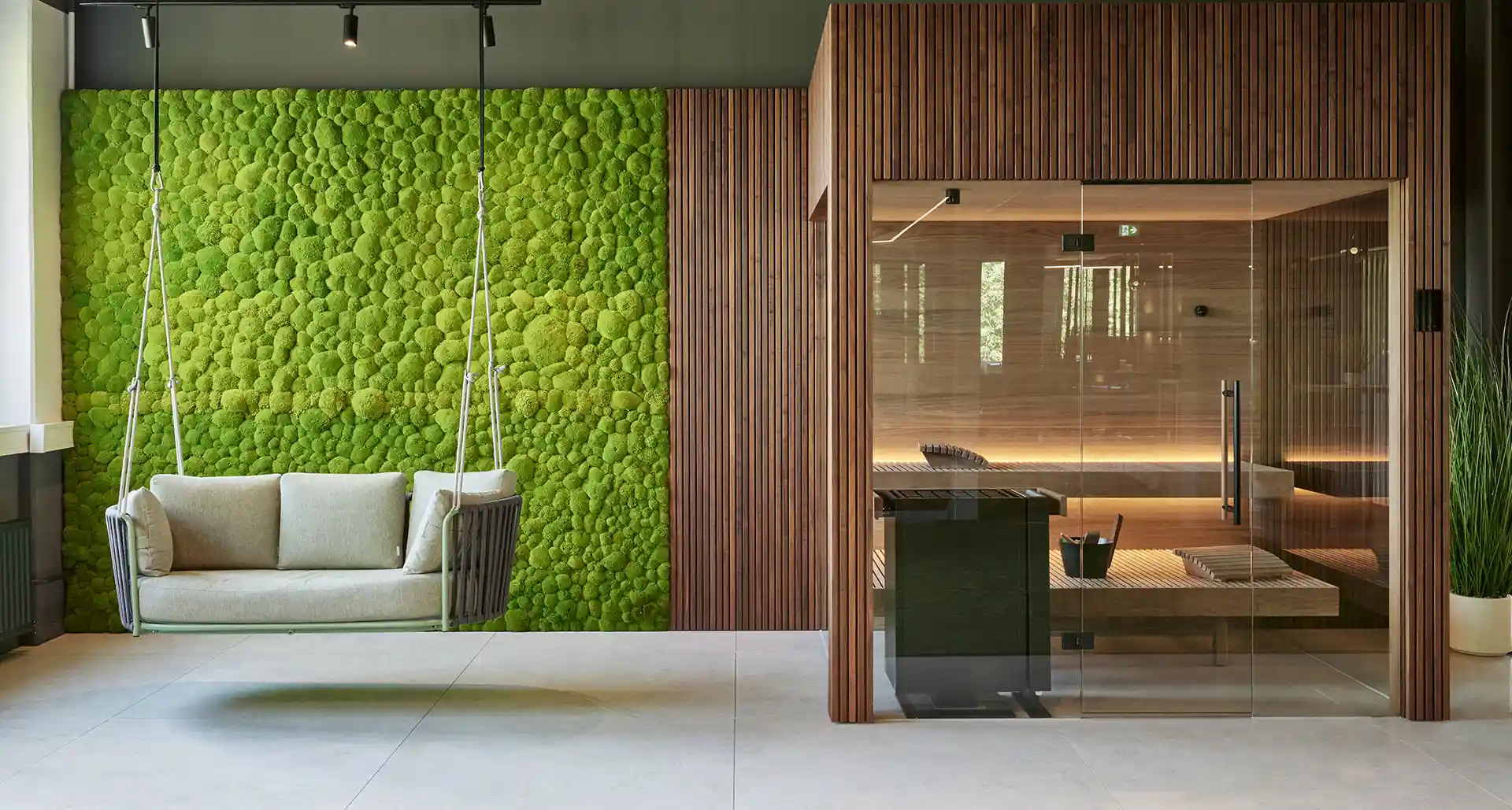Moderner Wellnessbereich mit einer großen grünen Mooswand, gemütlicher Hängesitzbank und verglaster Sauna, sorgt für eine natürliche und entspannende Atmosphäre.